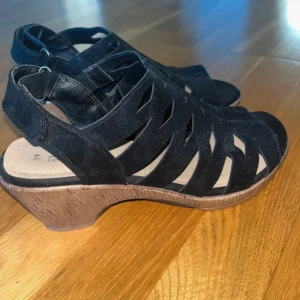 Svarta sandaler med klack från Alley - Säljer ett par svarta sandaler från Alley med snygga utskurna detaljer och öppen tå. Skorna har en bekväm klack i trälook och justerbar rem baktill. Perfekta för varmare dagar och ger en stilren touch till outfiten.