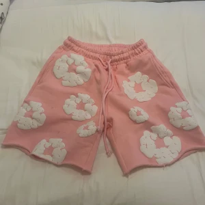 Rosa shorts med vita mönster från Denim Tears - Säljer ett par rosa shorts från Denim Tears med unika vita blommönster över hela plagget. Shortsen har elastisk midja med snörning och en bakficka. Perfekta för en avslappnad och trendig stil.