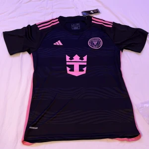 Inter Miami CF fotbollströja från Adidas - Säljer en svart Inter Miami CF fotbollströja från Adidas med rosa detaljer och klubbmärke på bröstet. Tröjan har korta ärmar, rosa ränder på axlarna och ett stort rosa tryck på framsidan. Perfekt för fotbollsfans dock storleken är lite större skulle säga M.