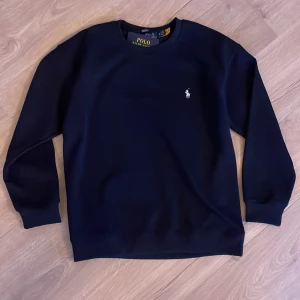 Marinblå sweatshirt från Polo Ralph Lauren - Säljer en klassisk marinblå sweatshirt från Polo Ralph Lauren med rund halsringning och broderad vit logga på bröstet. Tröjan har långa ärmar och ribbade muddar vid ärmslut och nederkant. Perfekt för en stilren och avslappnad look. 🏖️