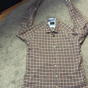 Rutig Peak Performance skjorta M - Snygg rutig skjorta från Peak Performance i storlek M. Skjortan har klassisk krage, knäppning framtill och långa ärmar. Färgerna går i brunt, blått och vitt, vilket ger en stilren och tidlös look. Perfekt för dig som gillar en avslappnad men ändå uppklädd stil.