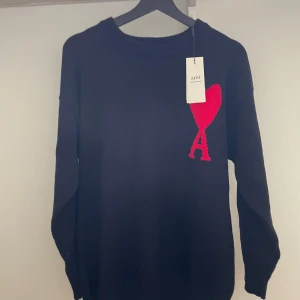 Svart stickad tröja från Ami Paris - Svart stickad tröja från Ami Paris med deras ikoniska röda hjärta och A-logga broderad på bröstet. Tröjan har rund halsringning, ribbade muddar och lång ärm. Perfekt för dig som gillar stilrena plagg med en twist.