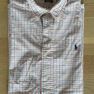 Rutig skjorta från Ralph Lauren M - Säljer en snygg rutig skjorta från Ralph Lauren i custom fit. Skjortan är vit med blå och orange rutor, har klassisk krage, knappar framtill och broderad logga på bröstet. Perfekt för dig som gillar stilrena och färgglada plagg.