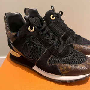 Louis Vuitton svarta sneakers med guld - Påse och box ingår | Snygga svarta Louis Vuitton sneakers med klassiskt monogrammönster i brunt och guld. Skorna har detaljer i mocka, mesh och skinn, samt guldfärgade metalldetaljer vid hälen. LV-logga på sidan och svarta snören. Perfekta för dig som vill sticka ut med lyxig streetstyle.
