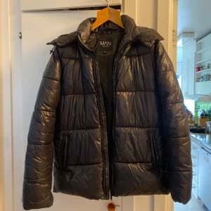Svart pufferjacka från Boohoo - Säljer en svart pufferjacka från boohoo MAN med huva och dragkedja. Jackan är quiltad och har en glansig finish, perfekt för kalla dagar. Den har två fickor med dragkedja och en hög krage som skyddar mot vinden. Enkel och stilren design som funkar till det mesta. Är från mansavdelingen men jag som tjej har haft mycket glädje av den. 