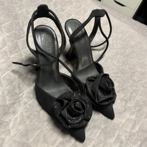 Svarta klackskor med rosdetalj - Säljer ett par svarta klackskor med spetsig tå och stor ros framtill. Skorna har smala remmar som går runt vristen och en elegant klack. Perfekta för dig som vill sticka ut med en unik detalj.