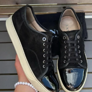 Svarta sneakers från Lanvin med lackad tå - Säljer ett par svarta sneakers från Lanvin med ovandel i mocka och snygg lackad tå. Skorna har vita sulor, svarta skosnören och diskreta sömdetaljer. Perfekta för dig som vill ha en stilren och lyxig look.