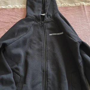 Svart hoodie från Better Bodies S - Svart hoodie från Better Bodies med dragkedja och huva. Klassisk och enkel design med logga på bröstet. Perfekt för gymmet eller chill dagar. Mjuk insida och två fickor framtill. Passar dig som gillar sportig och avslappnad stil.