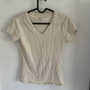 Beige v-ringad t-shirt i ekologisk bomull - Säljer en beige t-shirt från C i ekologisk bomull. T-shirten har v-ringning och korta ärmar, perfekt för en enkel och stilren look. Passar dig som gillar basic-plagg med skön passform.