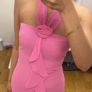 Rosa långklänning från adoore - Säljer en elegant rosa långklänning med halterneck och volangdetaljer framtill. Klänningen har en figurnära passform upptill och faller snyggt längs kroppen. Storlek 36 från adoore 