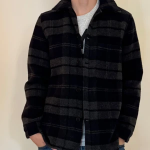 Rutig overshirt från A Days March - Snygg overshirt från A Days March i mörkblått och grått rutigt mönster. Jackan har klassisk krage, knäppning med svarta knappar och två bröstfickor. Tillverkad i mjuk ullblandning, perfekt för lager-på-lager under höst och vinter.