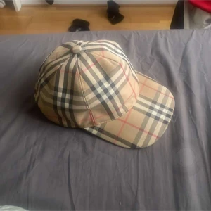 Burberry rutig keps i bomull - Snygg keps från Burberry i klassiskt beige rutigt mönster med inslag av svart, vitt och rött. Kepsen har böjd skärm och justerbar rem baktill. Tillverkad i bomull och perfekt för dig som vill ha en ikonisk accessoar med streetkänsla.