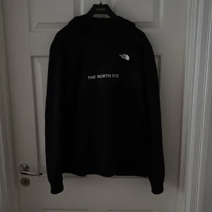 Svart hoodie från The North Face - Svart half-zip hoodie från The North Face med vit logga på bröstet och tryck över magen. Klassisk modell med huva och långa ärmar, perfekt för chill dagar eller när du vill ha en clean streetstyle-look.