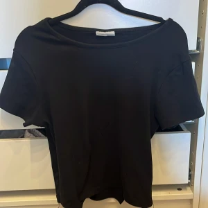 Svart basic T-shirt från Gina Tricot - En svart, enkel och stilren topp från Gina Tricot, basically basics-kollektionen. T-shirten har rund halsringning och korta ärmar, perfekt för en clean och avslappnad look. Gjord i mjuk bomull som känns skön mot huden.