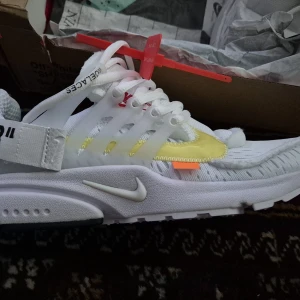 Nike Presto Off-White sneakers vit/gul - Nike Presto Off-White sneakers i vitt med detaljer i gult och orange. Skorna har en unik design med transparenta paneler, snörning och den klassiska Off-White zip-tagen i rött. Swoosh-logga på sidan och textdetaljer på remmen. Perfekt för dig som gillar statement-sneakers.