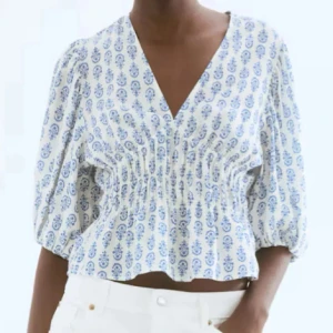Vit och blå blus med puffärm - Superfin blus i vitt med blått mönster, v-ringad och croppad passform. Blusen har puffiga trekvartsärmar och smockad midja som ger en snygg siluett. Perfekt för vår och sommar, till jeans eller kjol.