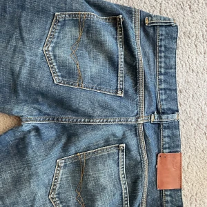 Blå jeans från Crocker - Snygga blå jeans från Crocker med slitna detaljer och råa kanter. Klassisk femficksmodell med orangea sömmar och knappstängning. Perfekt för dig som gillar en avslappnad och cool stil. Materialet är robust denim och shortsen har normal passform.