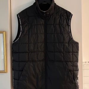 Svart dunväst från Calvin Klein Jeans - Svart quiltad dunväst från Calvin Klein Jeans med hög krage och dragkedja framtill. Diskret logga på ryggen och logoband längs ärmöppningarna.
