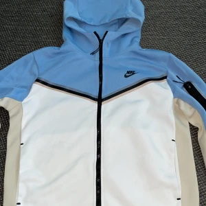 Nike vindjacka med huva, blå/vit - Nike vindjacka i storlek L passar M med snygg färgblock-design i ljusblått, vitt och beige. Jackan har huva, hel dragkedja framtill och en svart Nike-logga på bröstet. Materialet är polyester och jackan har långärmade ärmar samt en praktisk ficka med dragkedja på ena ärmen.