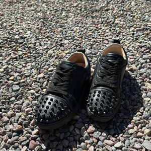 Svarta Louboutin sneakers med nitar - Snygga svarta sneakers från Christian Louboutin i skinn med ikoniska röda sulor. Skorna har svarta nitar över tån och klassisk snörning. Perfekta för dig som vill sticka ut med en edgy och lyxig vibe.