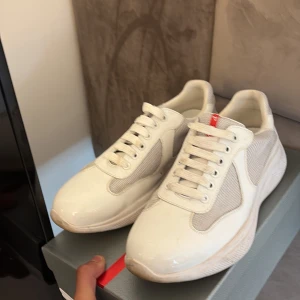  Prada cups - Snygga vita sneakers från Prada med meshpaneler och detaljer i blankt skinn. Klassisk design med rund tå, snörning och diskret röd logga på plösen och innersulan. Perfekta för dig som gillar stilrena och exklusiva sneakers.