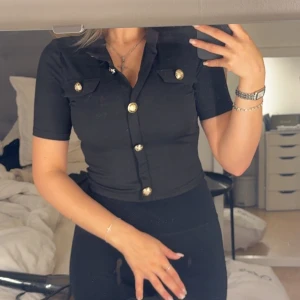 Svart croppad tröja med guldknappar - Snygg svart croppad tröja med korta ärmar och markerad krage. Toppen har dekorativa guldknappar framtill och på de två bröstfickorna. Ribbad struktur i tyget ger en extra touch. Perfekt för dig som gillar en stilren men ändå trendig look.