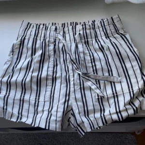 Randiga shorts med snörning från H&M - Snygga vita shorts från H&M med svarta vertikala ränder och bekväm snörning i midjan. Tillverkade i 100% bomull för en luftig känsla, perfekta för varma dagar. Elastisk midja och avslappnad passform gör dem superbekväma och lätta att styla. Inte använt någon gång så skicket är jättebra, pris kan diskuteras.