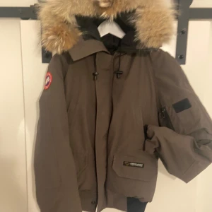 Brun dunjacka Canada Goose XS - Säljer en brun Canada Goose dunjacka med stor fluffig päls på huvan och klassisk logga på ärmen. Jackan har flera fickor, dragkedja och snörning vid huvan. Perfekt för kalla vinterdagar och riktigt snygg!! Jackan kan mycket väl vara äkta men även en kop1a men är osäker, skriv om ni har frågor!