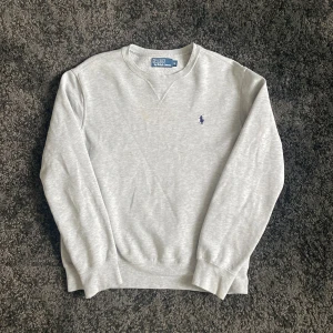 Grå Ralph Lauren sweatshirt - Klassisk grå sweatshirt från Polo Ralph Lauren. Tröjan har en fläck på bild 3 och en väldigt liten som sitter på vänster arm(bild 5). Annars är tröjan i perfekt skick! Hojta till om du har någon fråga!