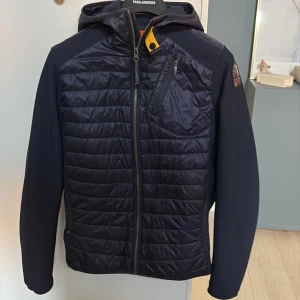 Mörkblå Parajumper cardigan - Snygg mörkblå cardigan från Parajumpers med huva och coolt badge på ärmen. Jackan har quiltad front, dragkedja hela vägen och en bröstficka med dragkedja. Perfekt för dig som vill ha en sportig och stilren look. Passar S-M Intressekoll! 