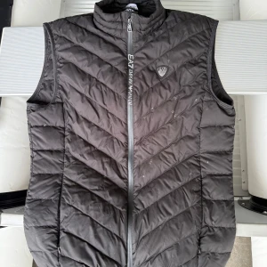Svart dunväst från EA7 Emporio Armani - Snygg svart dunväst från EA7 Emporio Armani med quiltad design och hög krage. Västen har dragkedja framtill med logga och ett EA7-märke på bröstet. Perfekt att slänga över en hoodie för en sportig look.