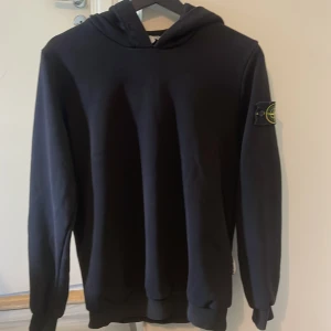 Svart hoodie från Stone Island - Svart hoodie från Stone Island med klassisk logga på vänster ärm. Tröjan har huva, långa ärmar och ribbade muddar. Perfekt för dig som gillar streetwear och vill ha en stilren look. Materialet är mjukt och skönt, troligtvis bomull. Snören följer med såklart 😊😊