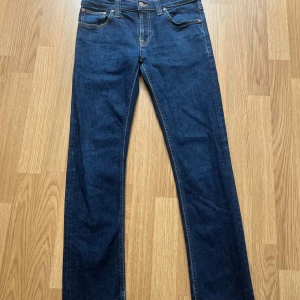 Mörkblå jeans från Nudie Jeans, 28/32 - Snygga mörkblå jeans från Nudie Jeans med klassisk femficksdesign och kontrastsömmar. Modellen har rak passform och är tillverkad i slitstarkt bomullsjeans. Perfekta för dig som gillar stilrena och tidlösa jeans med diskreta detaljer.