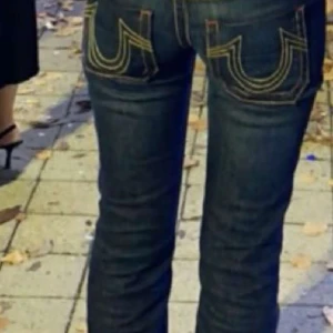 Mörkblå bootcut jeans från True Religion - Säljer ett par mörkblå jeans från True Religion med ikoniska kontrastsömmar och stora U-formade fickor bak. Modellen har låg midja och bootcut-ben, vilket ger en snygg siluett. Jeansen är tillverkade i klassiskt denim och har detaljer som knappar och dragkedja framtill.