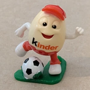 Unik samlarfigur från Kinder som spelar fotboll - Unik samlarfigur från Kinder med röd keps. Figuren är gjord av plast och står på en grön platta av plast med en klassisk svartvit fotboll.