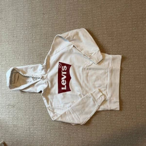 Vit hoodie från Levi's med röd logga - Säljer en vit hoodie från Levi's i storlek XXS. Tröjan har en stor röd Levi's-logga på bröstet, huva med snörning och en klassisk magficka. Mjuk och skön bomullskvalitet, perfekt för chill dagar. Långärmad och med ribbade muddar vid ärmslut och nederkant.