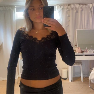 Svart långärmad topp med spets - Svart långärmad topp med snygg v-ringning och spetsdetaljer längs halsringningen. Modellen är figurnära och croppad, vilket ger en trendig look. Perfekt att styla med jeans eller kjol för en cool vibe.