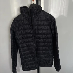 Svart dunjacka från J.Lindeberg - Snygg svart dunjacka från J.Lindeberg med quiltad design och huva. Jackan har dragkedja framtill, två sidofickor och diskret logga på ärmen. Perfekt för kyliga dagar och enkel att matcha med allt. Lätt och smidig att bära med sig.