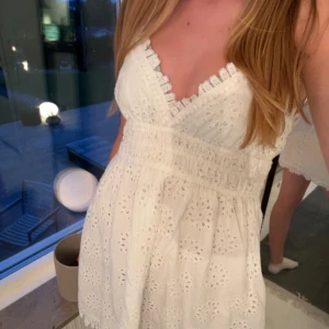 Vit broderad sommarklänning  - Superfin vit kortklänning med broderade blommor, v-ringning och spetsdetaljer. Smockad midja och tunna axelband ger en söt och luftig känsla. Perfekt för varma dagar och somriga vibes!