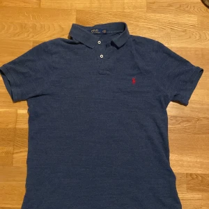 Blå Polo Ralph Lauren t-shirt slim fit - Snygg blå t-shirt från Polo Ralph Lauren i slim fit-modell. Klassisk pikékrage med två knappar och röd broderad logga på bröstet. Tillverkad i mjuk bomull som känns skön mot huden. Perfekt för dig som gillar stilren och enkel design.
