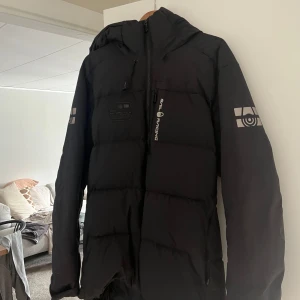 Svart Sail Racing dunjacka med huva - Säljer en svart dunjacka från Sail Racing med huva och coola loggor på ärmarna. Jackan har dragkedja framtill och är quiltad för extra värme. Perfekt för dig som vill ha en stilren och sportig look under kalla dagar. Nypris 4500kr , den ser oanvänd ut och inga synliga skador på jackan 