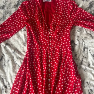 Röd blommig långklänning från Adoore - Superfin röd långklänning från Adoore med vita små blommor och v-ringad hals. Klänningen har långa ärmar och knappar hela vägen framtill. Perfekt för dig som gillar romantisk stil och vill sticka ut med färg och mönster. Stl 34 men passar 36