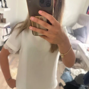 Vit t-shirt med spetsdetalj - Säljer en fräsch vit t-shirt med korta ärmar och snygga spetsdetaljer vid ärmsluten. T-shirten har en clean och enkel design som känns både stilren och modern. Perfekt för dig som gillar en feminin touch på din outfit.