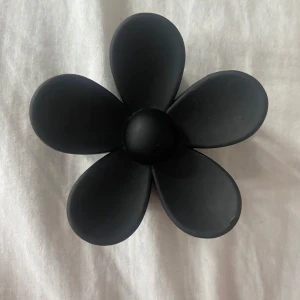 Svart blommig klämma i plast - Snygg svart hår klämma med stor blomma i matt plast. Blomman har fem rundade kronblad och ger en cool statement-look. Perfekt accessoar för att lyfta din outfit med en unik touch.