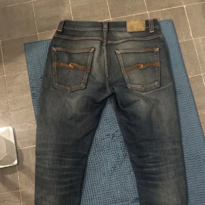 Mörkblå Nudie Jeans slim fit - Snygga mörkblå jeans från Nudie Jeans med klassisk femficksdesign och coola orangea sömmar på bakfickorna. Modellen är slim fit med normal passning och raka ben. Jeansen är tillverkade i slitstark denim av bomull och har subtila slitningar för en schysst look.