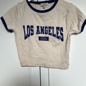 Beige croppad t-shirt Pull&Bear - Snygg croppad t-shirt från asos (pull and bear) i beige med mörkblå detaljer vid hals och ärmslut. Tryck med 'Los Angeles 1991' i blått framtill. 