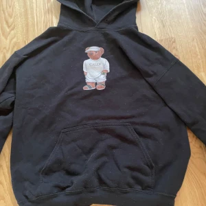 Svart hoodie med Cally-björn - Svart hoodie från Cally med en söt björn i tennisoutfit tryckt på bröstet. Tröjan har huva, stor magficka och långa ärmar. Perfekt för chill dagar och streetstyle. Materialet känns mjukt och skönt mot huden.