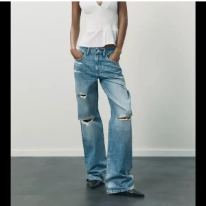 Trendiga zara jeans - Säljer ett par ljusblå jeans med breda ben och snygga slitningar på både fram- och baksida. Jeansen har klassisk femficksdesign, hög midja och är tillverkade i bomull. Perfekta för en avslappnad och trendig look. Använda 1 gång
