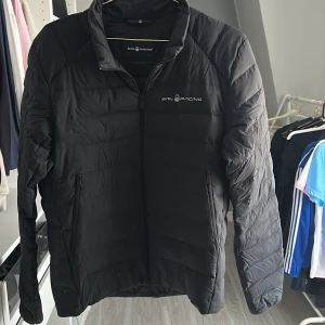 Svart pufferjacka från Sail Racing - Svart pufferjacka från Sail Racing med diskret logga på bröstet och ryggen. Jackan har dragkedja framtill, två sidofickor med dragkedja och quiltad design. Perfekt för kyliga dagar och har en clean, sportig look som funkar till det mesta.