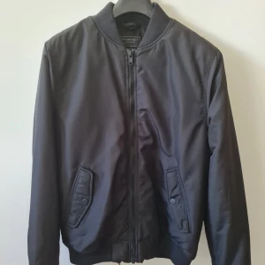 Svart bomberjacka från H&M strl M - Svart bomberjacka från H&M i storlek M. Jackan har klassisk ribbad krage, inne ficka, muddar och nederkant, samt dragkedja framtill. Två fickor med knappstängning och quiltat innerfoder. Lättfodrat jacka.